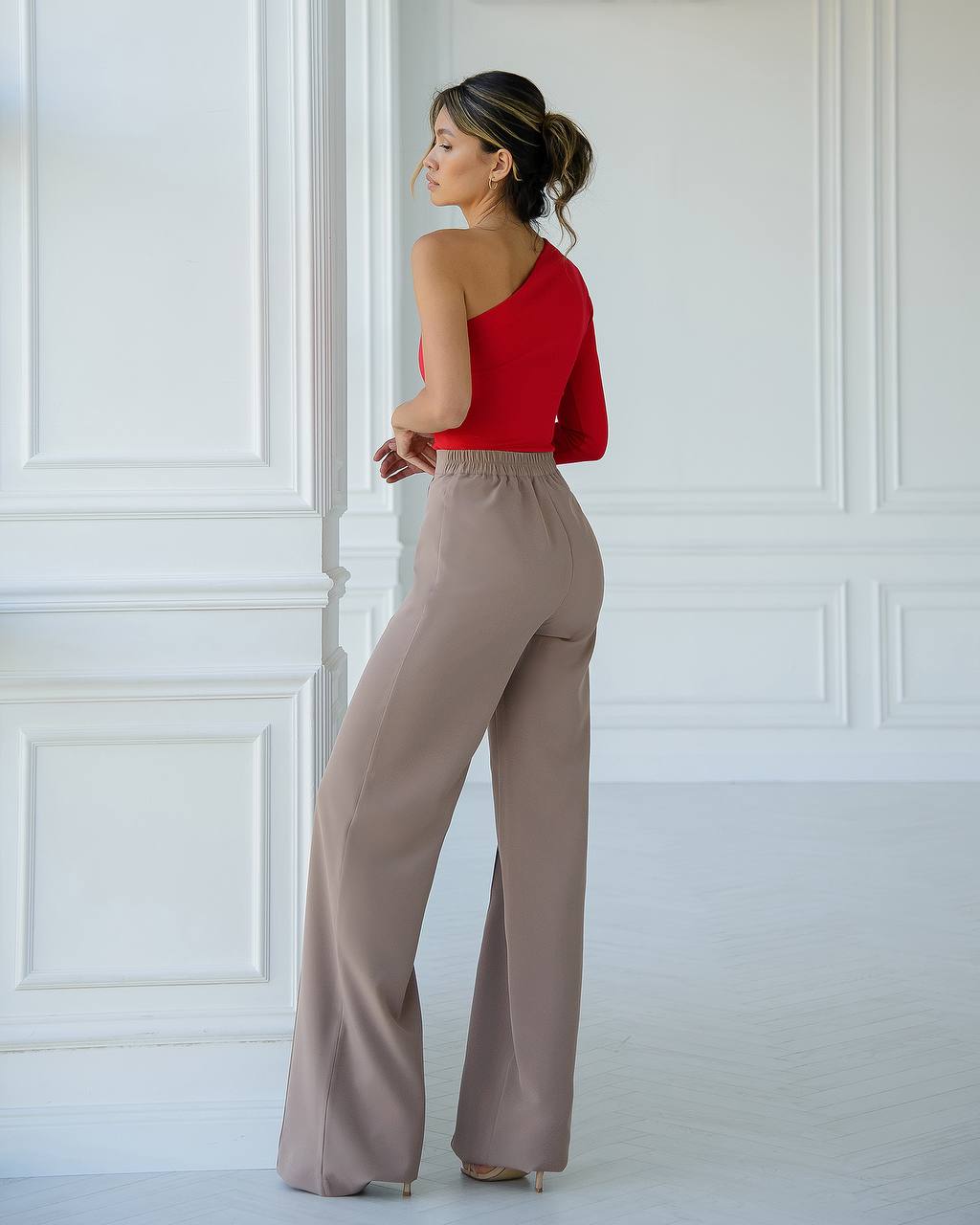 Woman wearing a red top and beige pants against a white paneled wall.