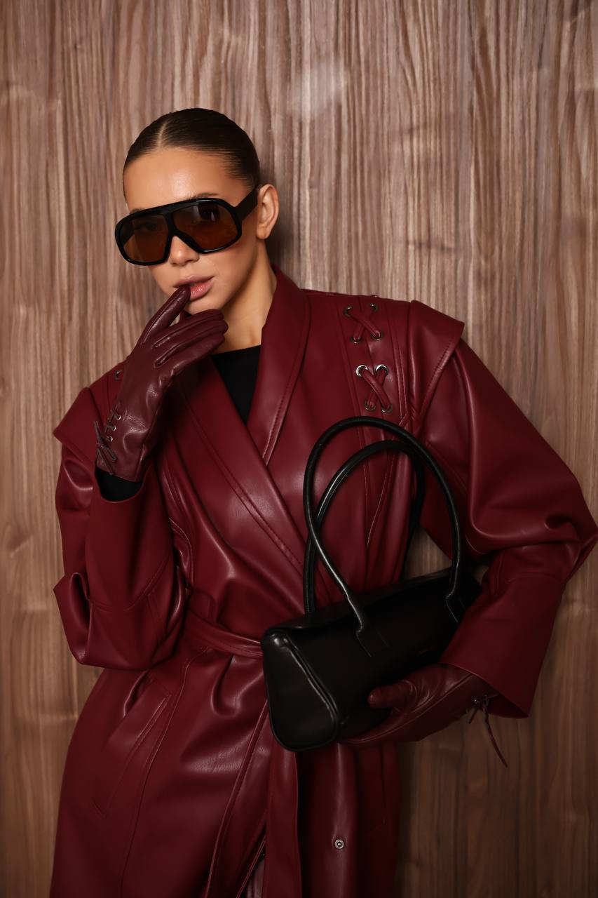 Person wearing a red leather coat and sunglasses, holding a black handbag against a wooden background