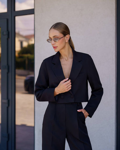 Woman wearing a black suit standing against a neutral wall.