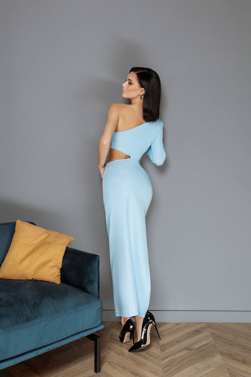 Woman in a light blue dress standing against a gray wall with a blue couch and yellow pillow.