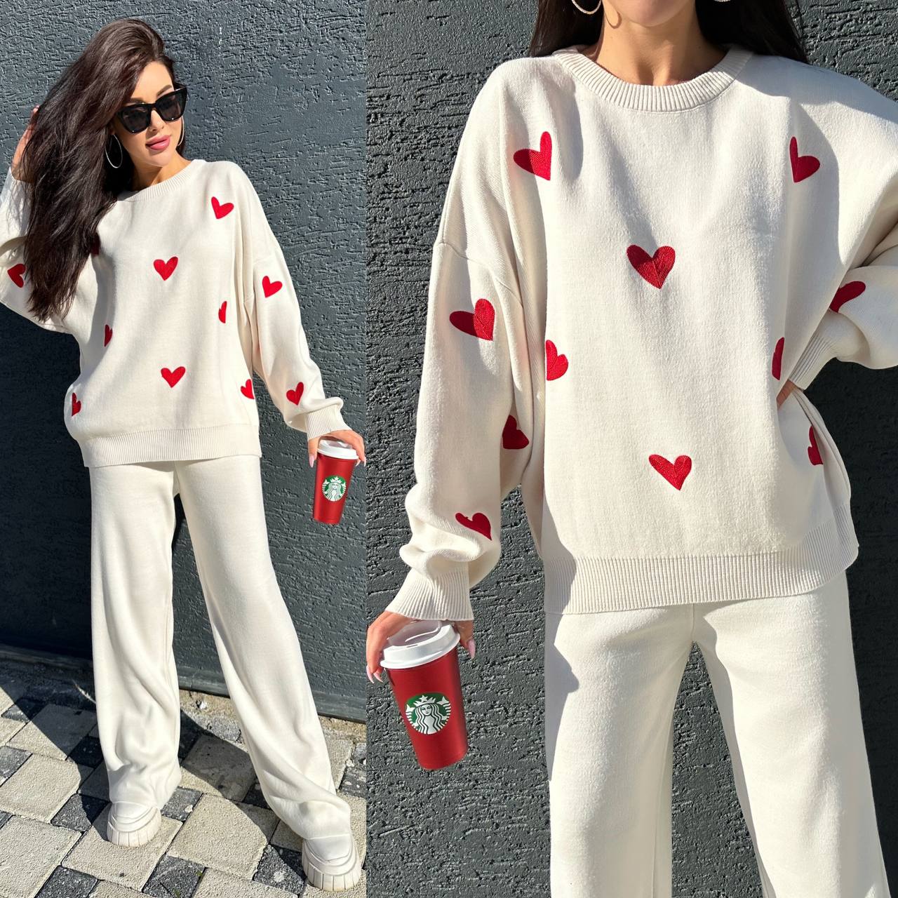 Woman wearing a white sweater with red heart patterns and white pants, holding a red Starbucks cup against a gray wall.