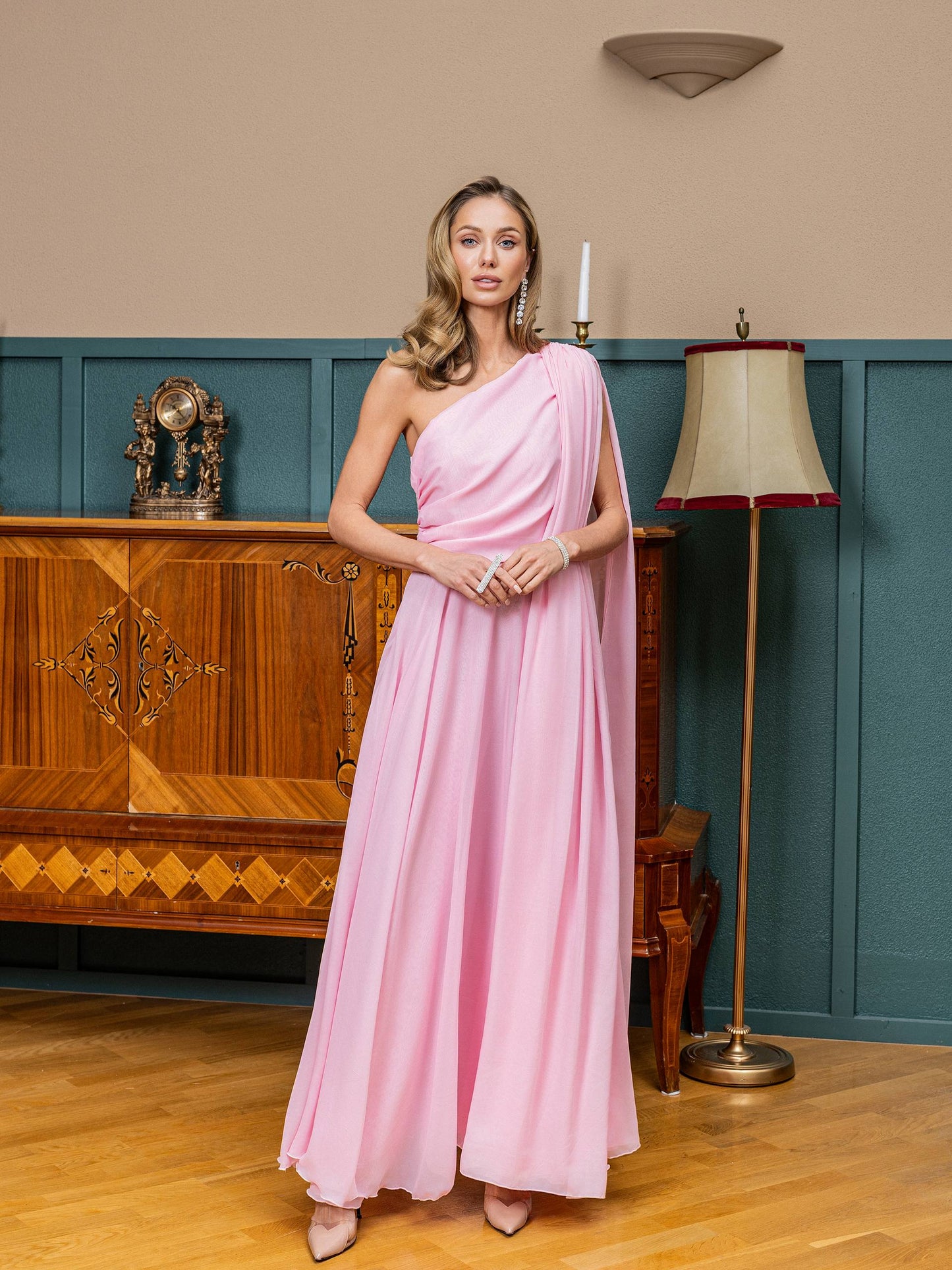 Woman in a pink one-shoulder dress standing in an elegant room with wooden furniture and a lamp.