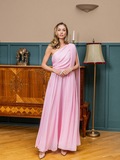 Woman in a pink one-shoulder dress standing in an elegant room with wooden furniture and a lamp.