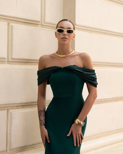 Woman wearing a green off-shoulder dress with gold accessories against a neutral wall.
