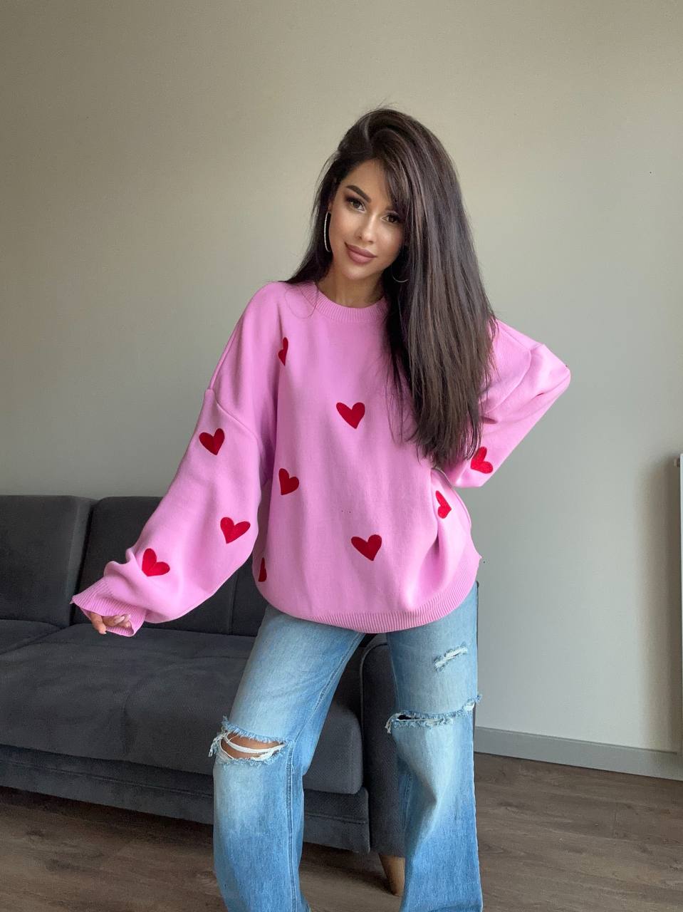 Woman wearing a pink sweater with red heart patterns and ripped jeans in a living room.