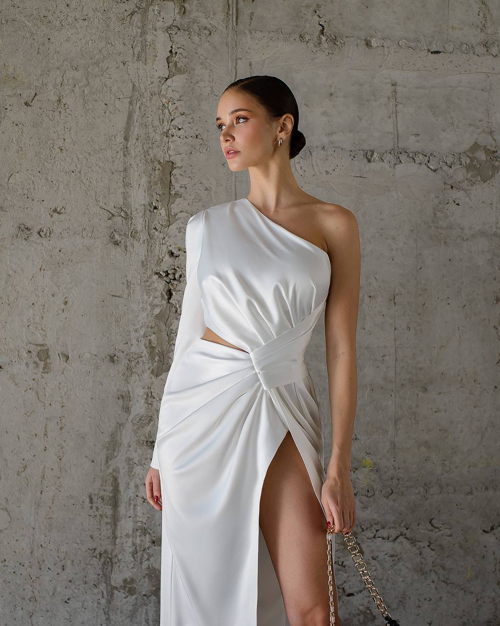 Woman wearing a white one-shoulder dress against a textured wall.