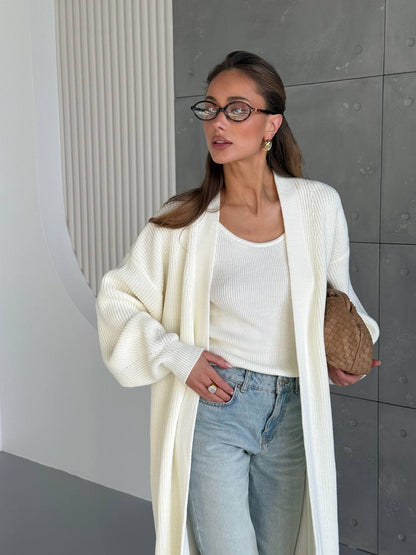 Woman wearing a white cardigan, white top, and light blue jeans against a gray wall.