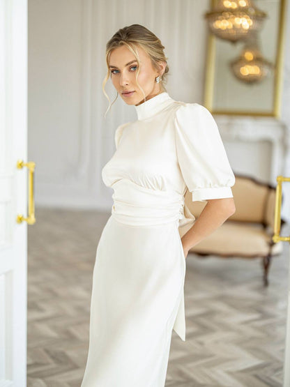 A woman wearing a white high-neck midi dress with puffed short sleeves and a gathered waist, standing in a bright, elegant room with a beige armchair and gold accents in the background.