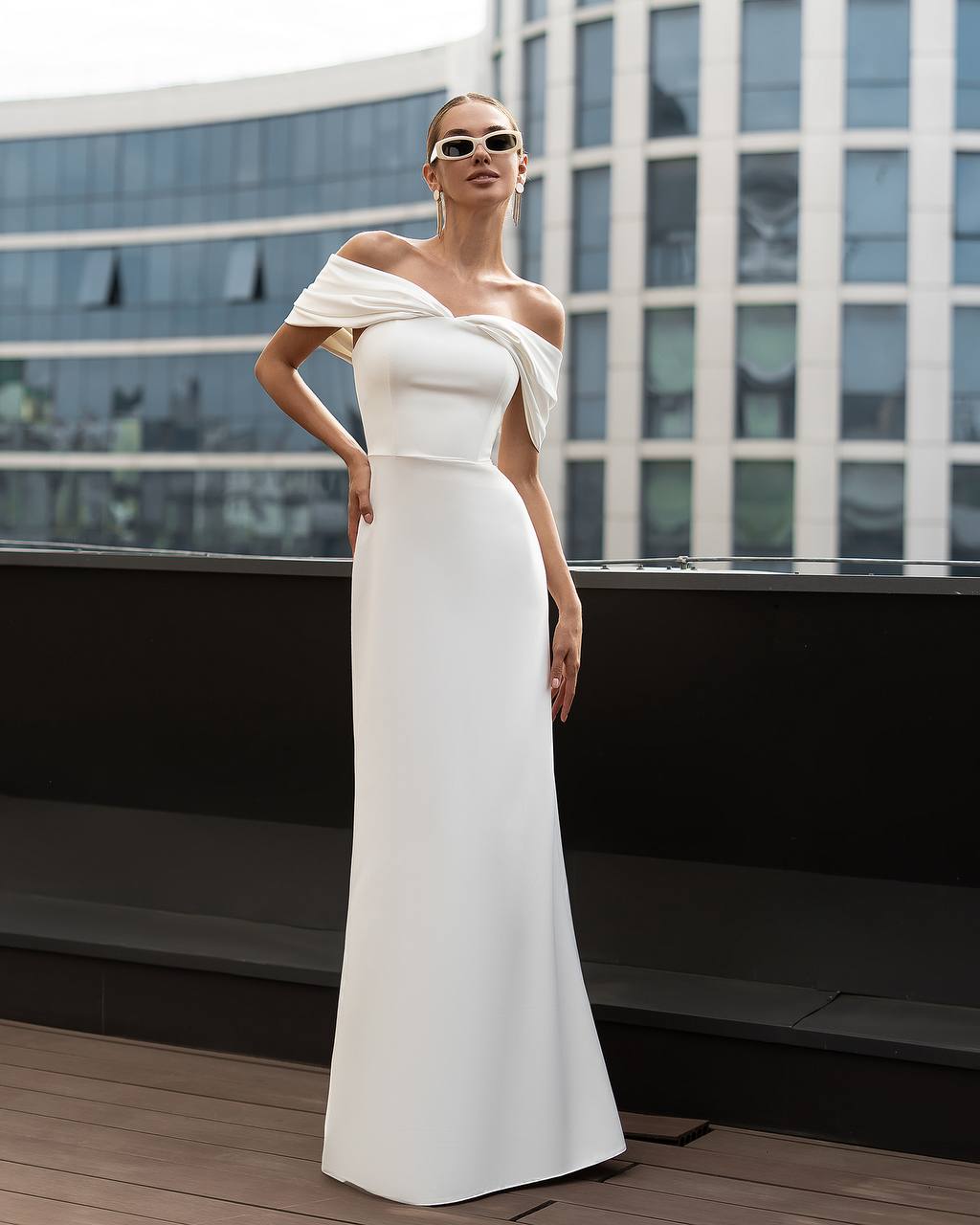 Woman in a white off-shoulder gown standing on a rooftop with modern building in the background