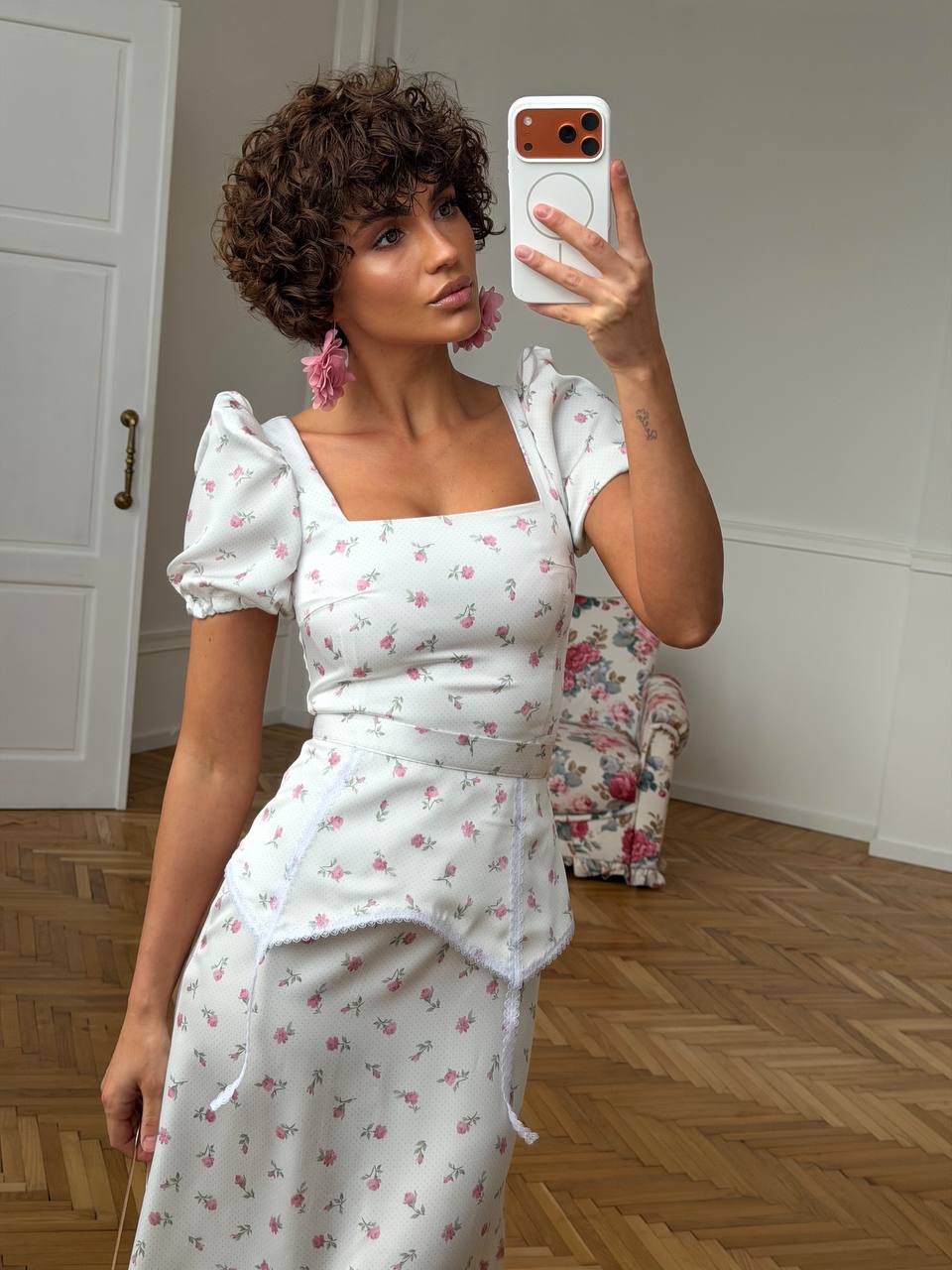Woman taking a mirror selfie wearing a white floral dress in a room with wooden flooring.