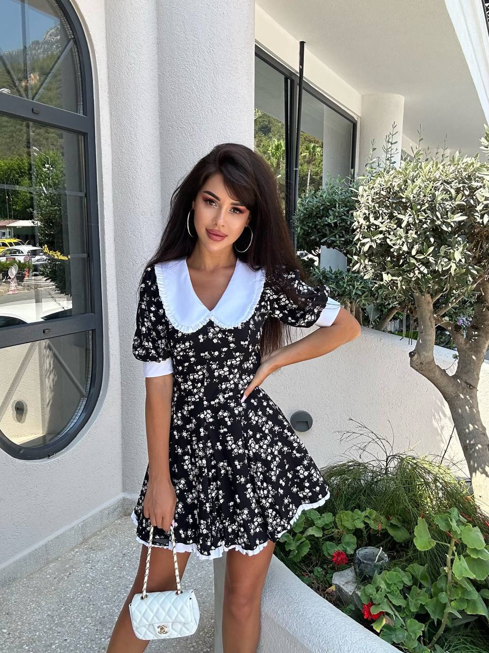 Woman in a black floral dress with white collar standing outdoors near a building.