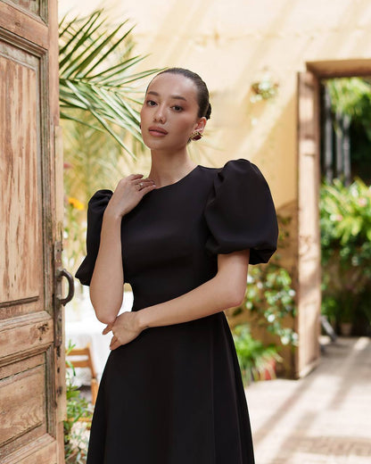 Woman in a black dress standing in a sunlit outdoor setting with plants and wooden doors.