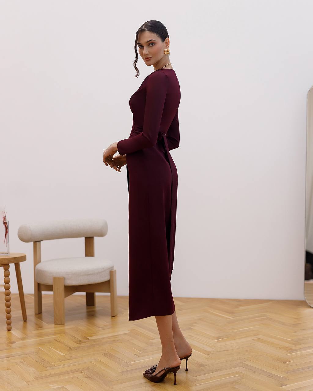 Woman in a burgundy dress standing in a room with wooden flooring and a white wall.