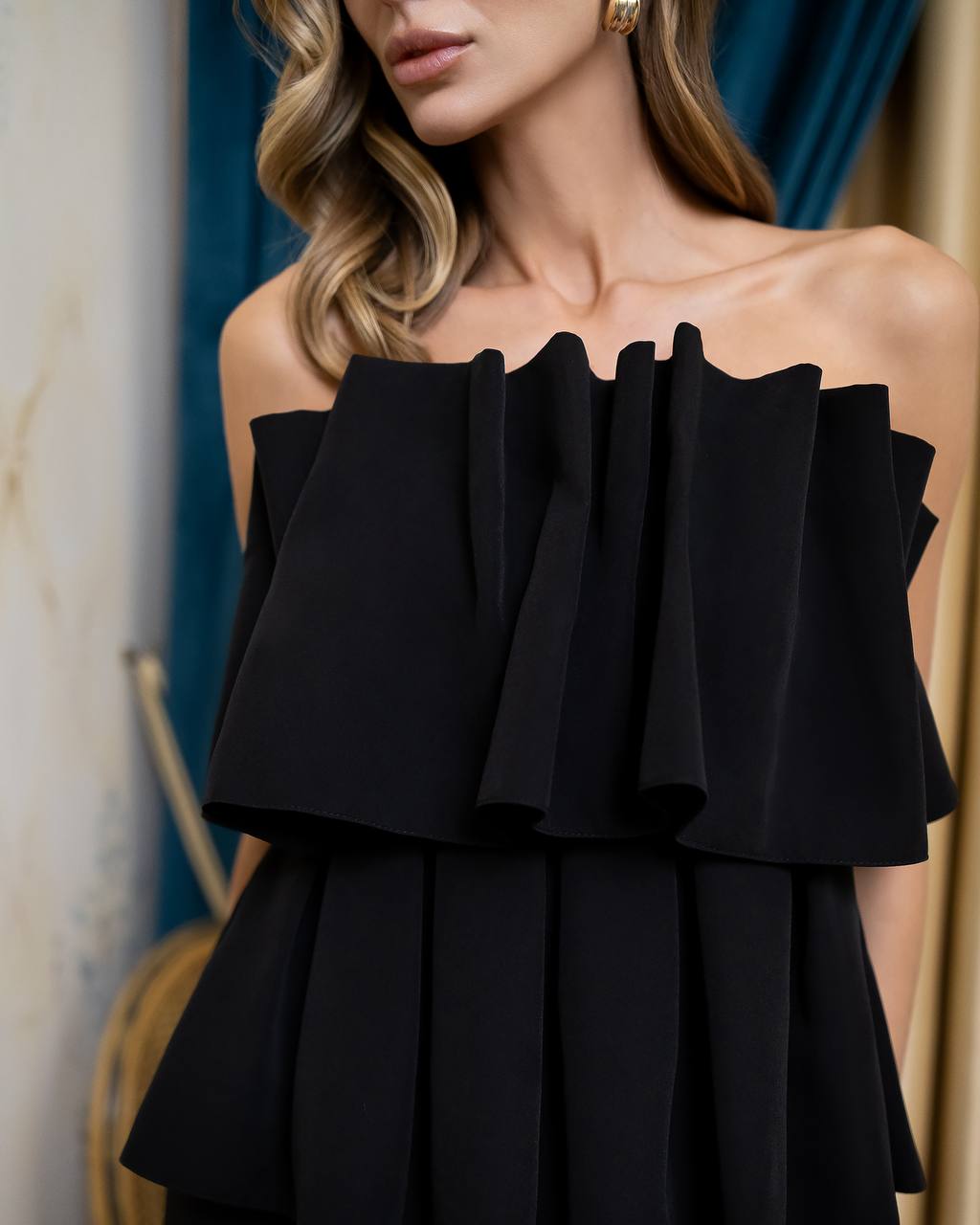 A close-up of a woman wearing a black off-the-shoulder dress with dramatic layered ruffles across the bodice and a pleated skirt, photographed indoors against blue and beige drapery.
