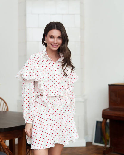 Woman wearing a white dress with red polka dots in a room with wooden furniture and a white curtain.