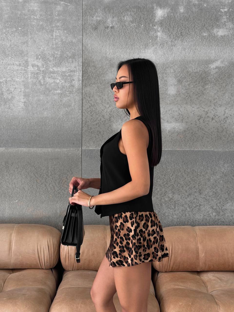 Woman in black sleeveless top and leopard print shorts standing against a gray wall with a beige couch.