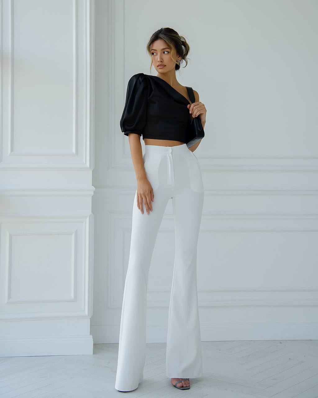 Woman wearing a black top and white pants against a white wall