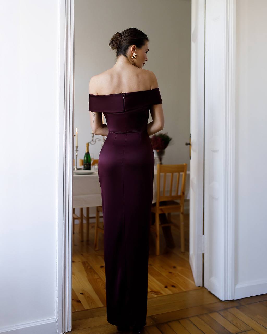 Woman in a dark purple off-shoulder gown standing in a room with wooden flooring and white walls.