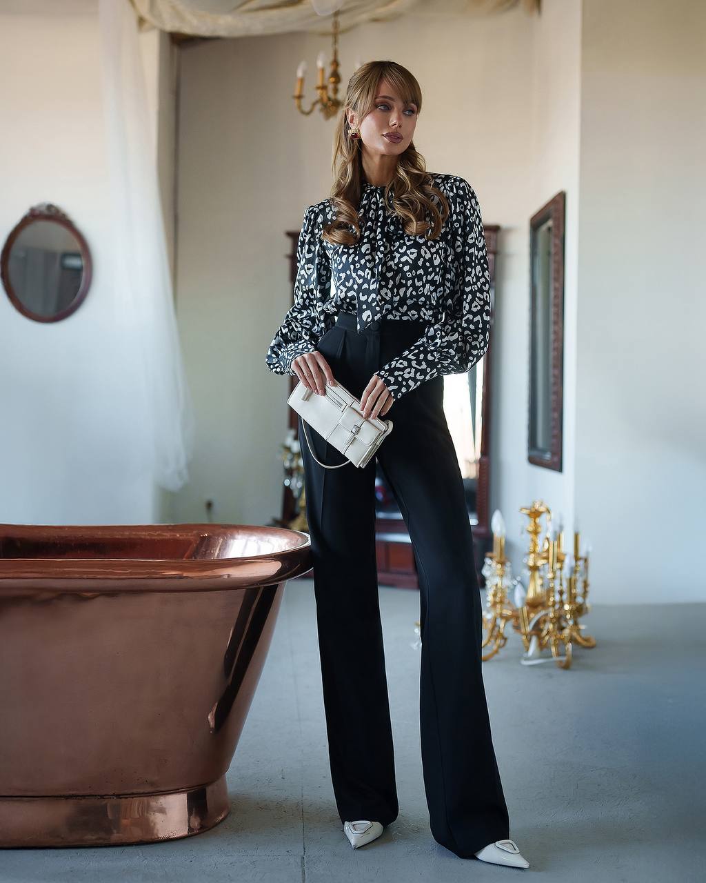 Woman in a patterned blouse and black pants standing in an elegant room with a copper bathtub.