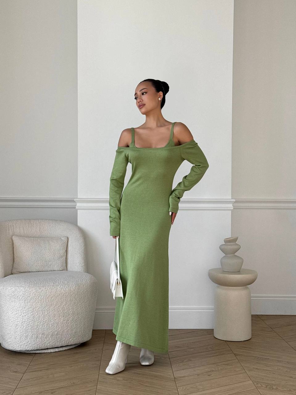 Woman in a green dress standing in a room with a white wall and beige chair.
