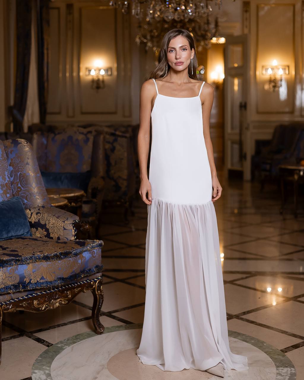 Woman in a white dress standing in an elegant room with chandeliers and ornate furniture.