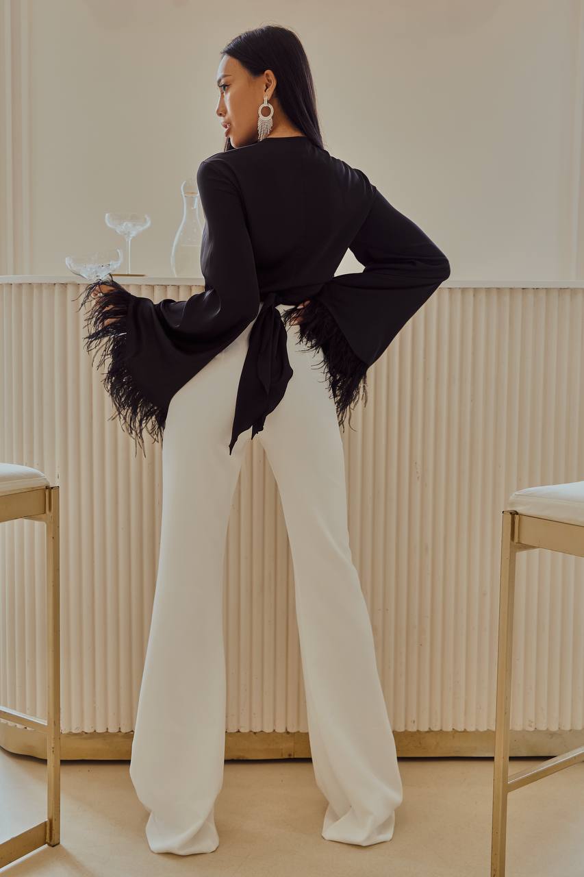 Woman in a black jacket with feather details and white pants standing in a room with beige walls and chairs.