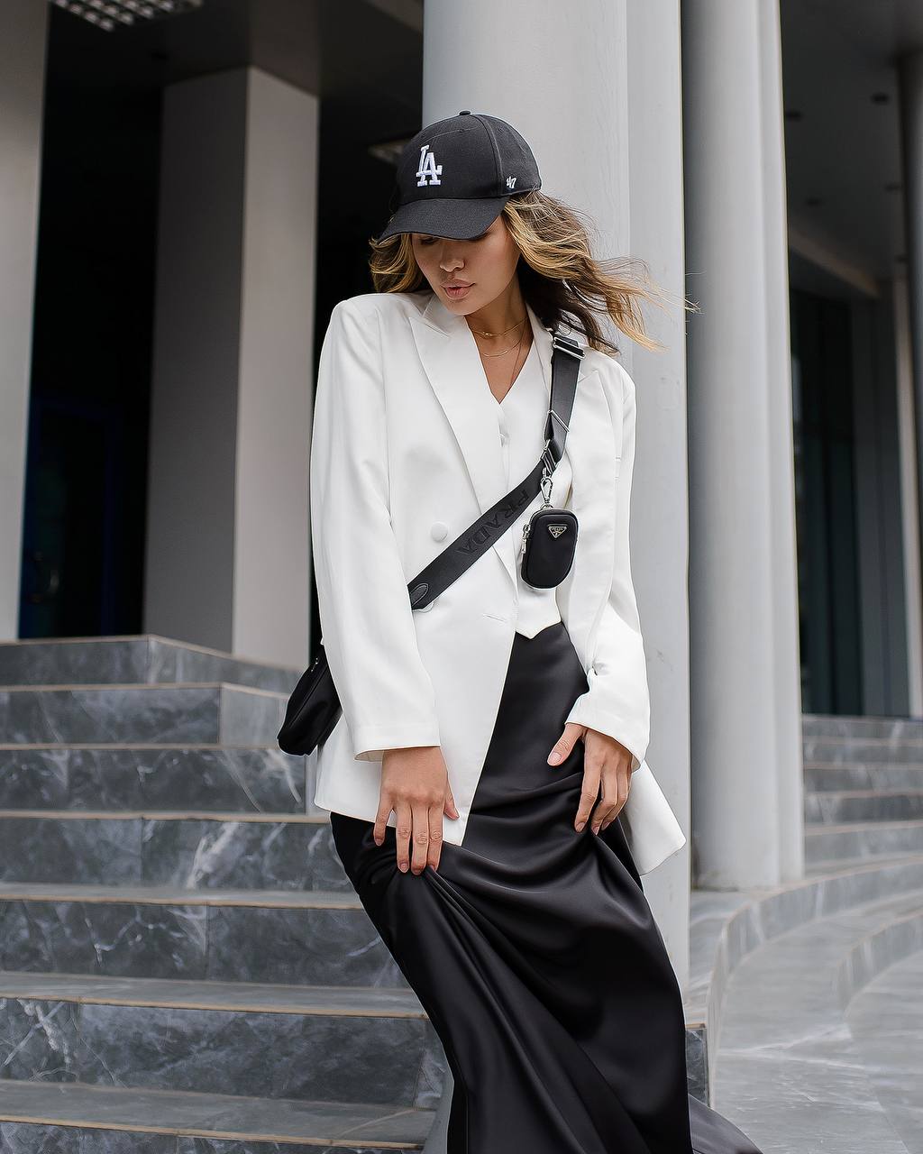 Woman in a white blazer and black skirt standing on steps with a black bag and cap.