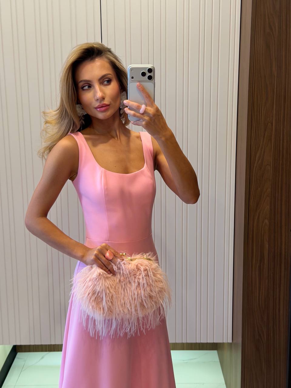 Woman in a pink dress holding a feathered clutch in front of a mirror.