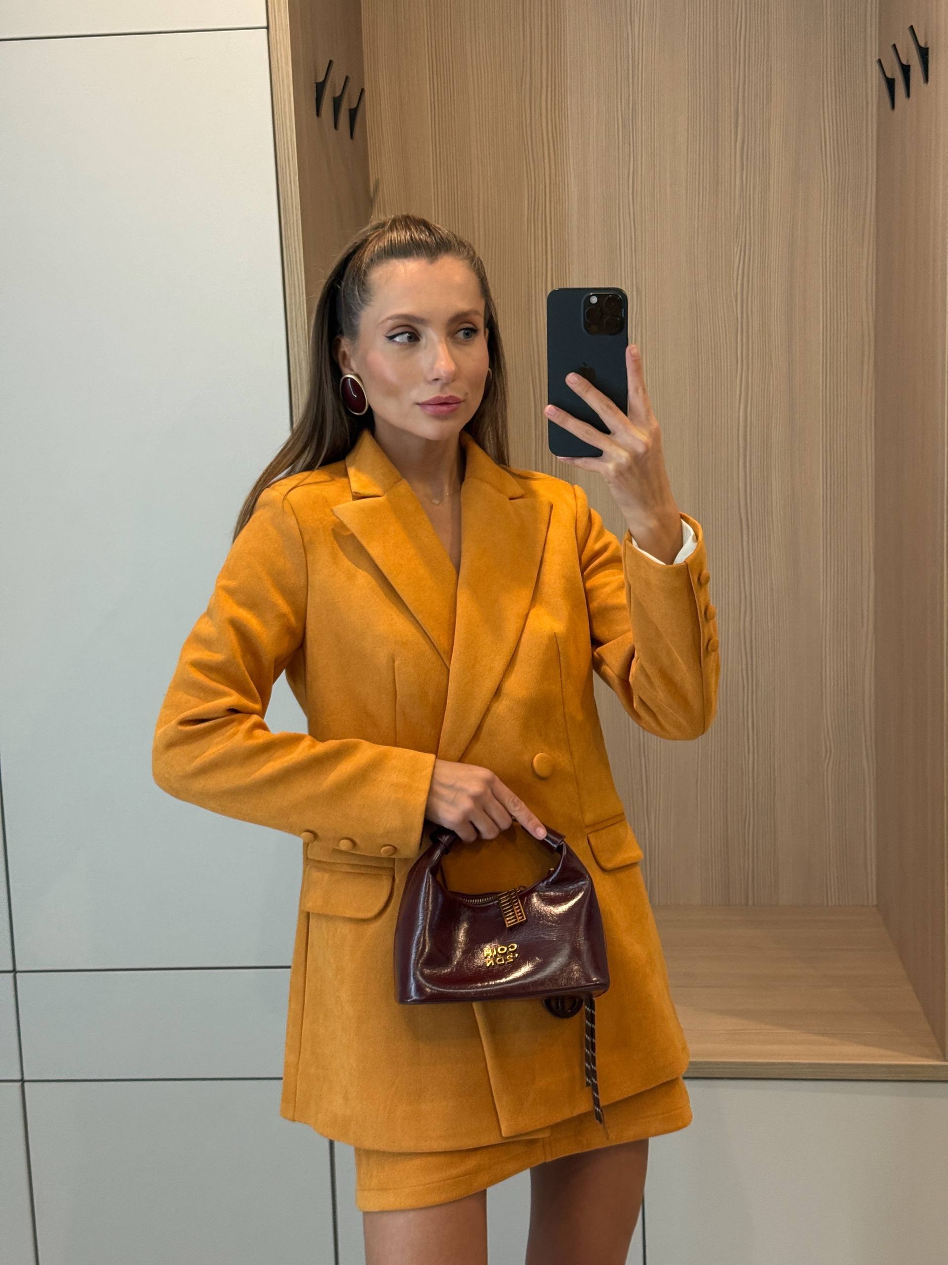 Woman taking a mirror selfie wearing a mustard yellow suit and holding a brown handbag.