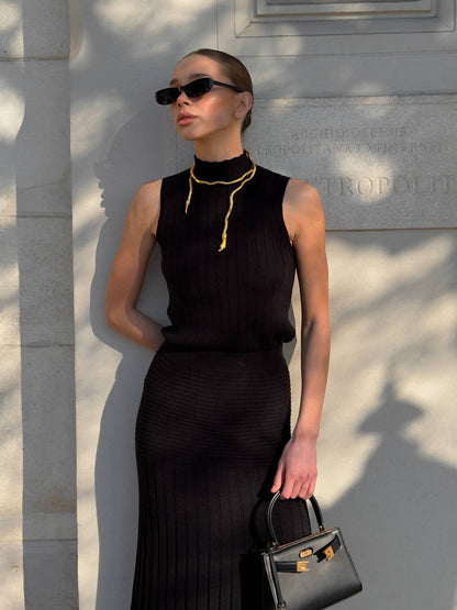 Woman in a black dress with a gold necklace, sunglasses, and a black handbag against a stone wall.