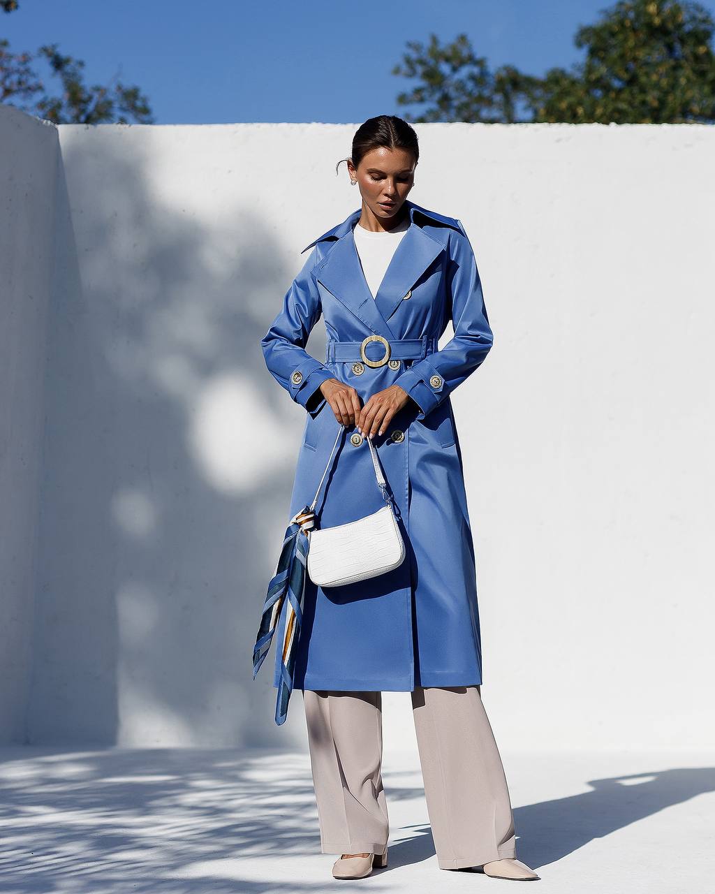 Woman wearing a blue trench coat holding a white handbag against a white wall with blue sky.
