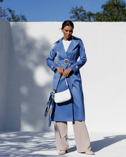 Woman wearing a blue trench coat holding a white handbag against a white wall with blue sky.