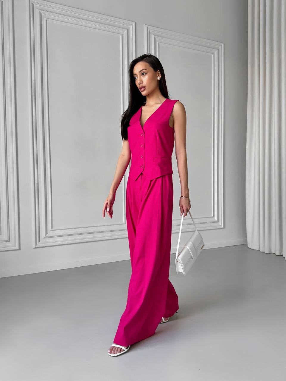 Woman in a bright pink jumpsuit holding a white clutch against a gray wall.