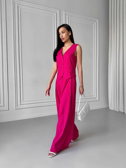 Woman in a bright pink jumpsuit holding a white clutch against a gray wall.