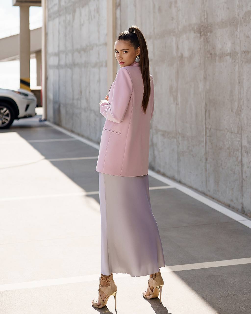 A woman stands in a concrete parking structure wearing a light pink blazer and a long lavender-to-white ombre skirt, paired with beige high-heeled sandals.