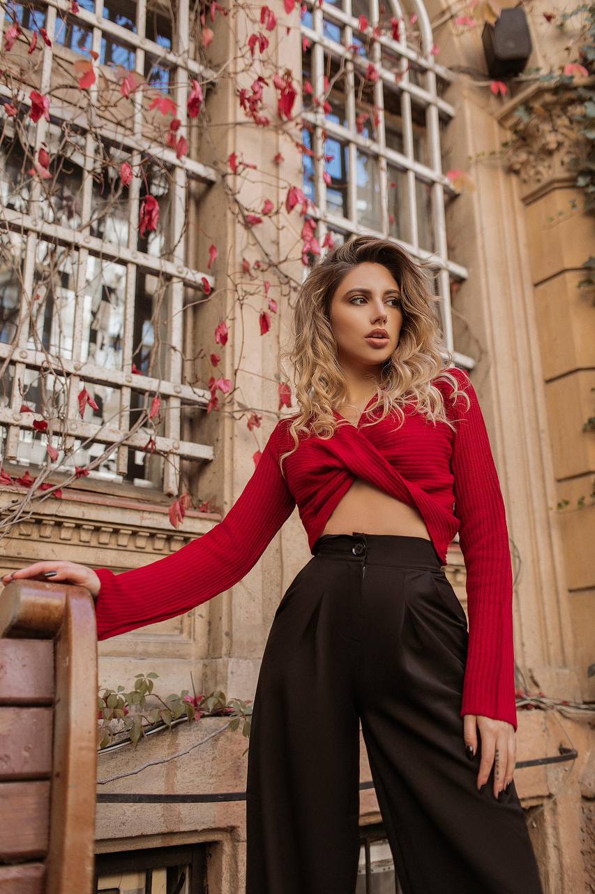 Woman in a red top and black pants standing against a decorative wall with plants.