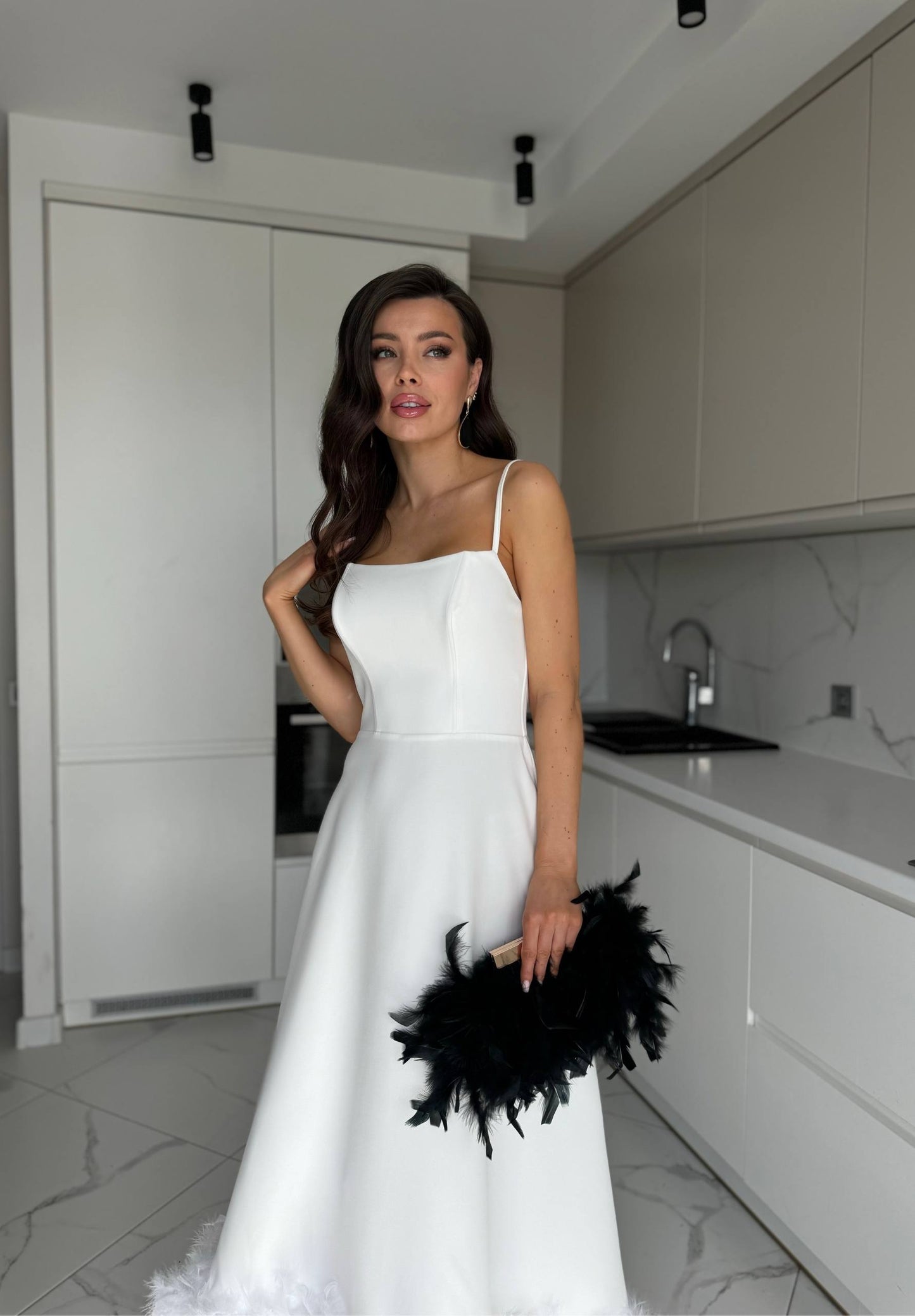 Woman in a white dress holding black feathers in a modern kitchen.