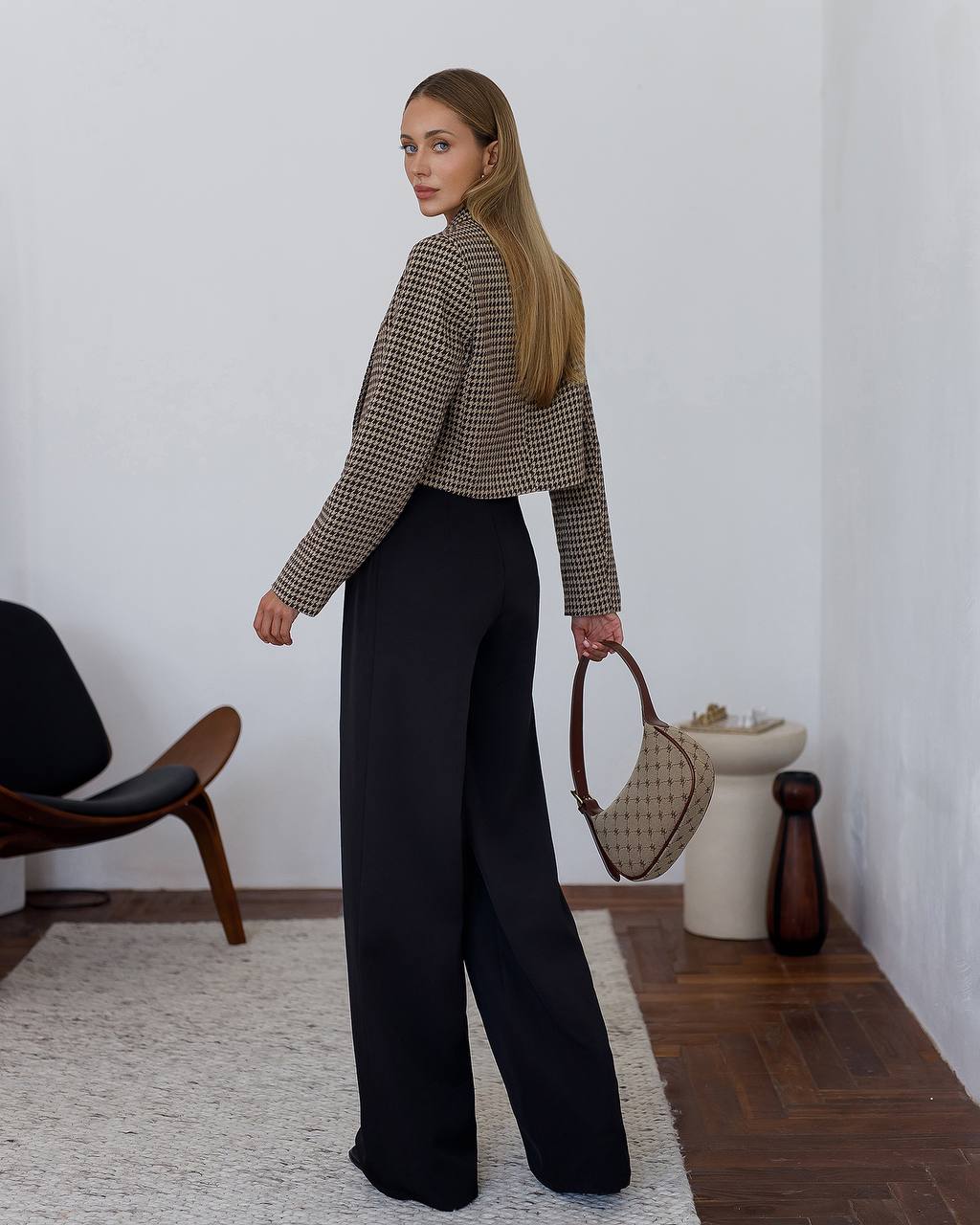 Woman in a checkered shirt and black pants holding a handbag in a room with minimal decor.