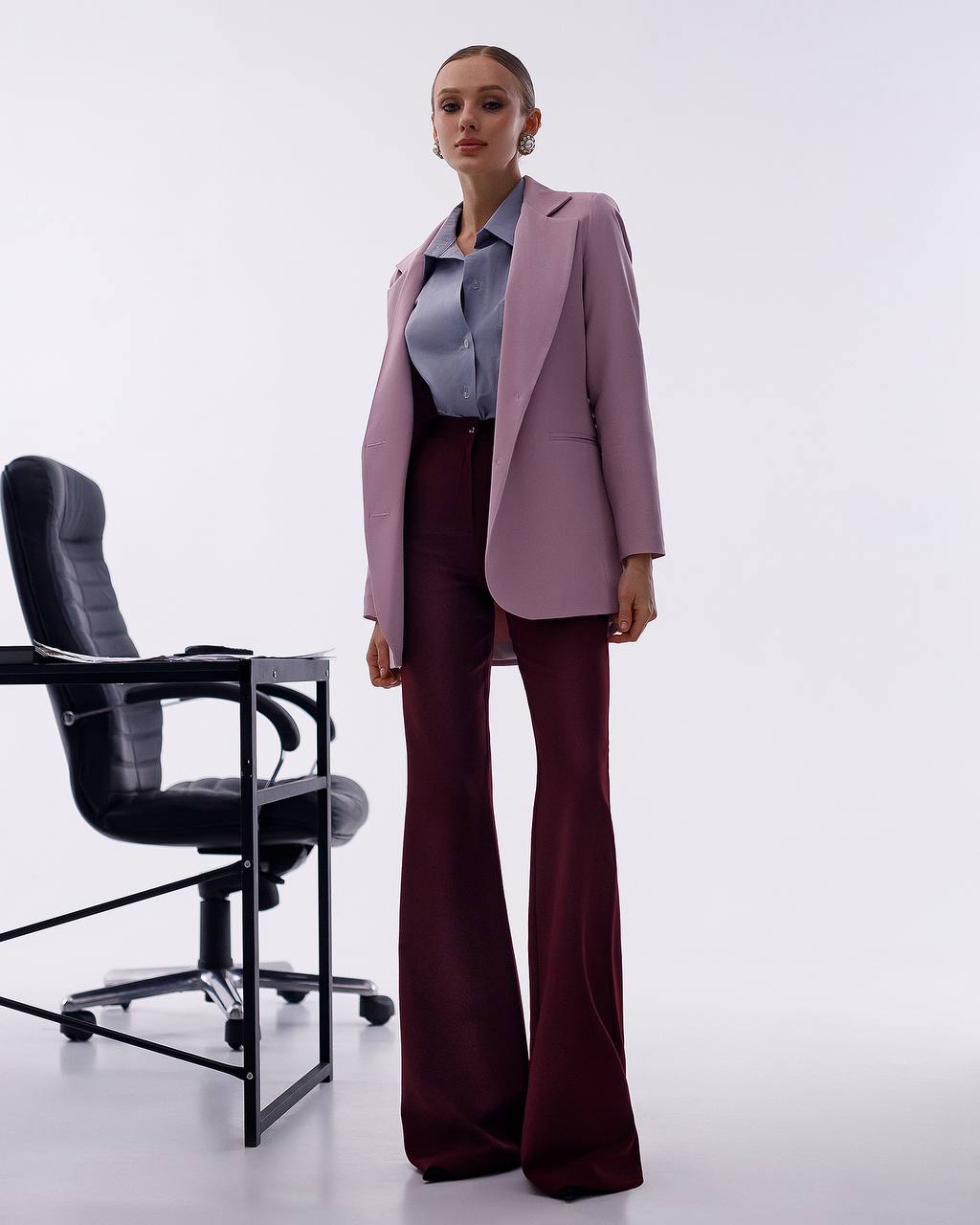 A woman models a two-piece pantsuit featuring a dusty lavender blazer and matching wide-leg trousers in a deep burgundy, styled with a light blue button-down shirt. She stands in a studio setting next to a black office chair against a plain white background.