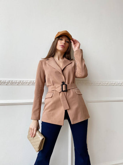 Woman wearing a beige trench coat with a belt, holding a beige clutch against a white wall.