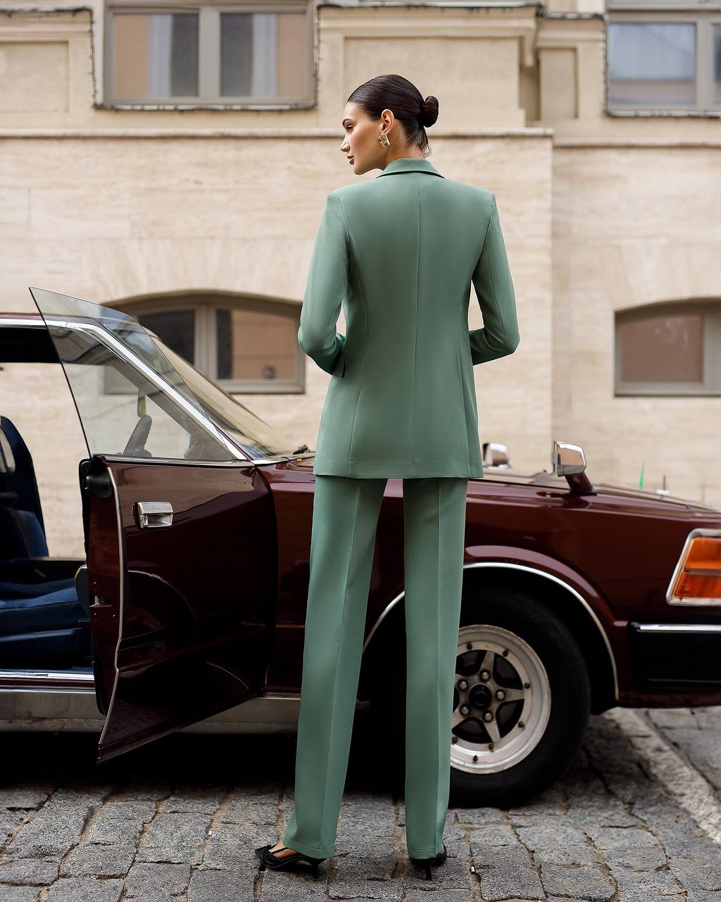 Woman in a green suit standing next to an old-fashioned car on a city street.