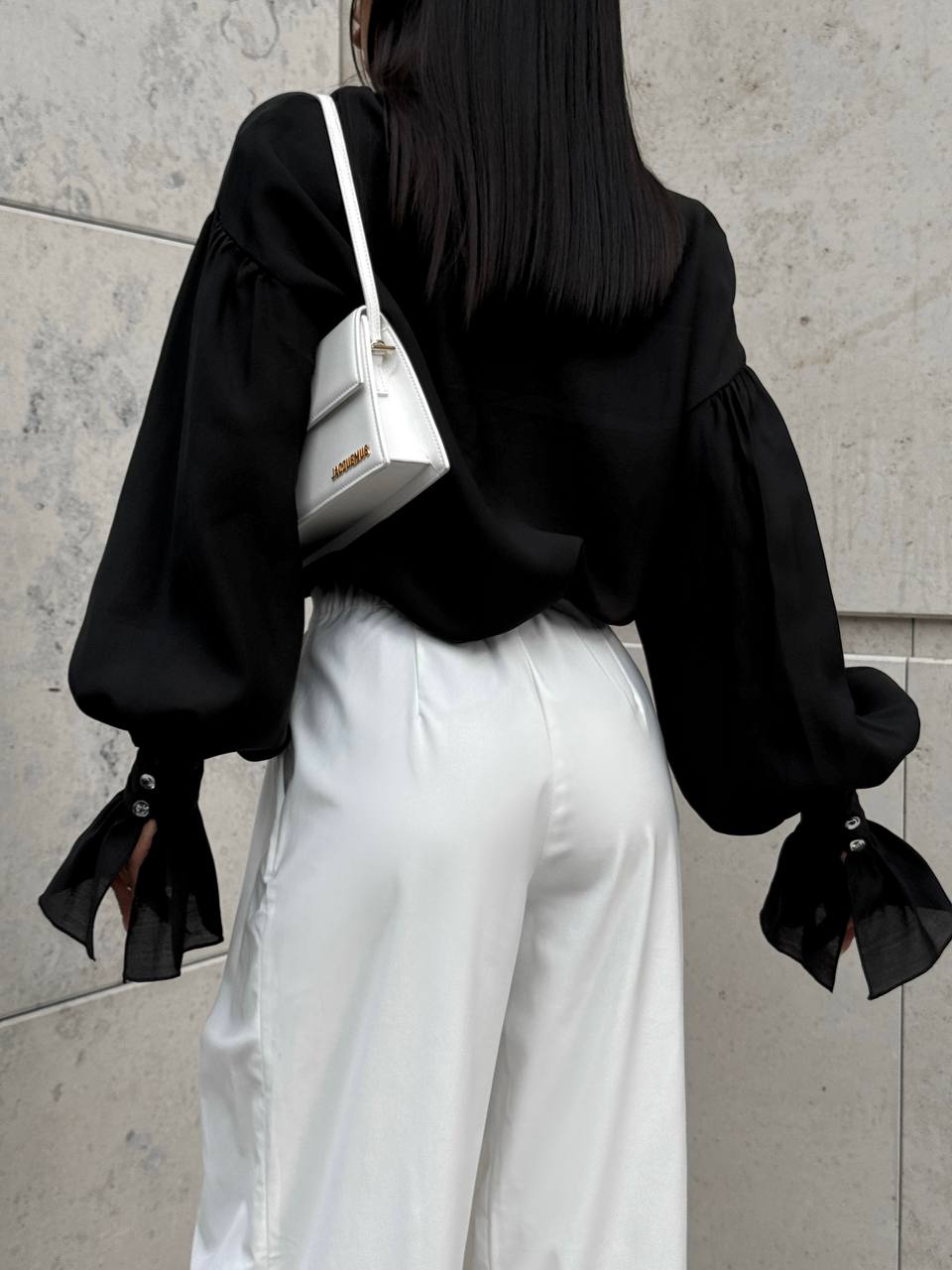 Person wearing a black blouse and white pants with a white handbag, standing against a light gray wall.