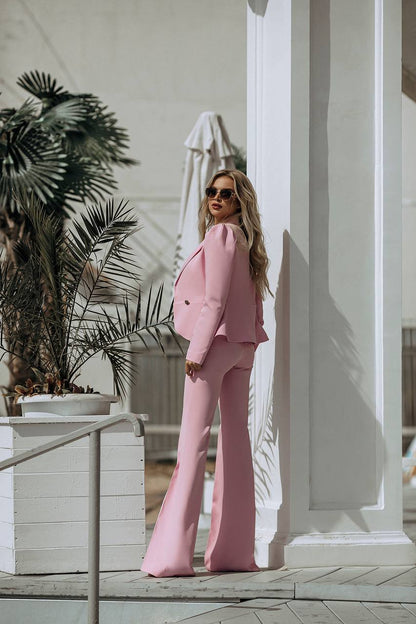 Woman in a pink suit standing outdoors with plants and a white building in the background