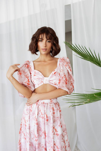 Woman wearing a pink floral dress standing against a white curtain with a plant in the foreground.