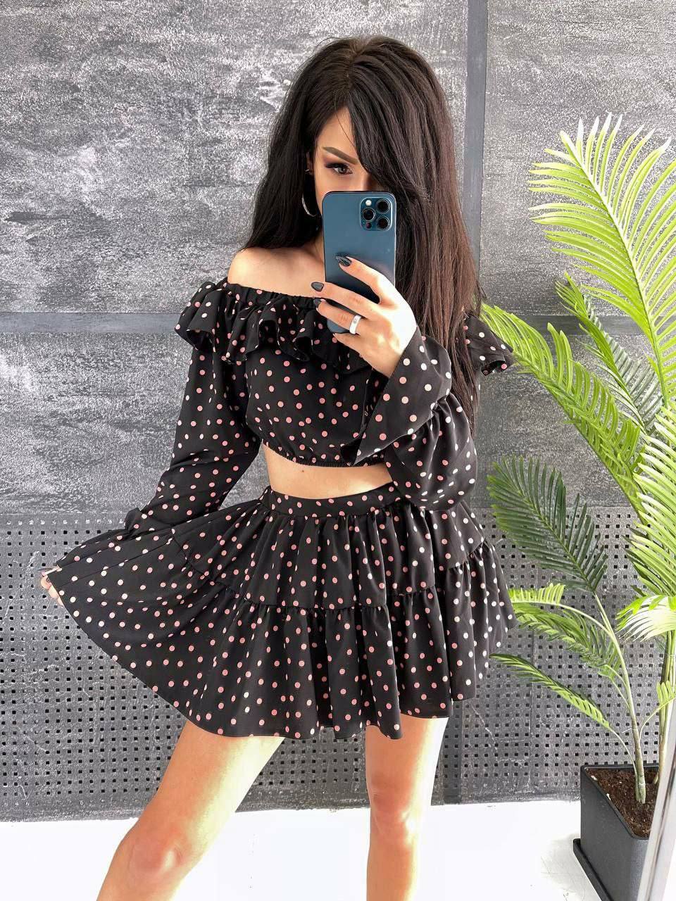 Woman taking a mirror selfie wearing a black polka dot dress against a gray wall with a plant.