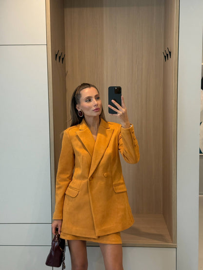 A woman takes a mirror selfie wearing a mustard yellow double-breasted blazer and matching mini skirt. She is standing in front of a light wood and white cabinet background, holding a dark burgundy handbag.