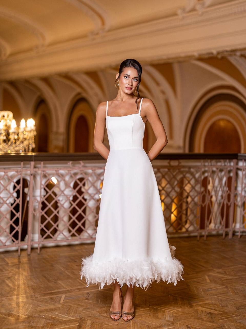 Woman in a white dress with feather details standing in an elegant indoor setting.