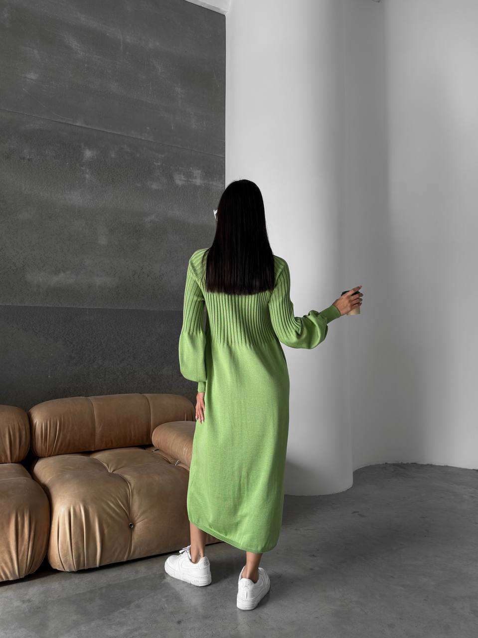 Person wearing a green dress standing in a room with a brown sofa and gray walls.