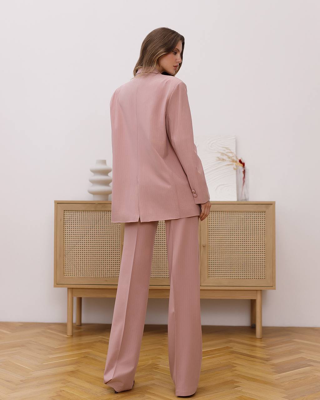 Woman wearing a pink suit standing in a room with wooden flooring and a light-colored wall.