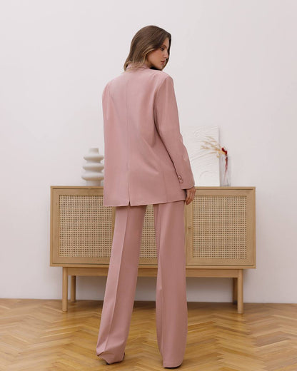 Woman wearing a pink suit standing in a room with wooden flooring and a light-colored wall.